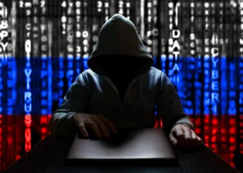 Unknown Russian hacker opens the lid of a laptop, sitting on the background of the Russian flag from a binary code.
