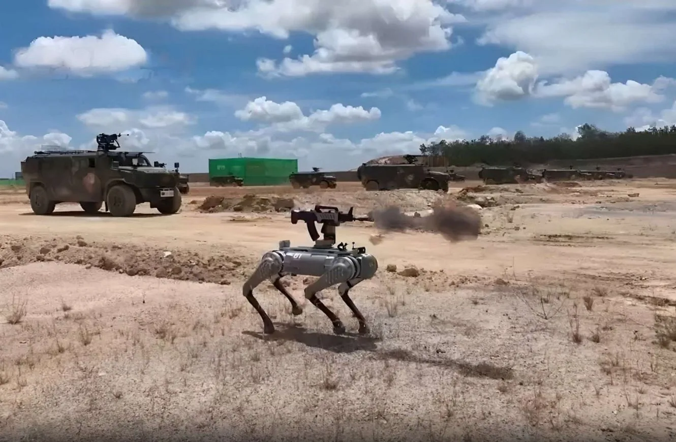 Un robot cuadrúpedo con montaje de arma realiza una demostración en campo abierto, junto a vehículos blindados en segundo plano.