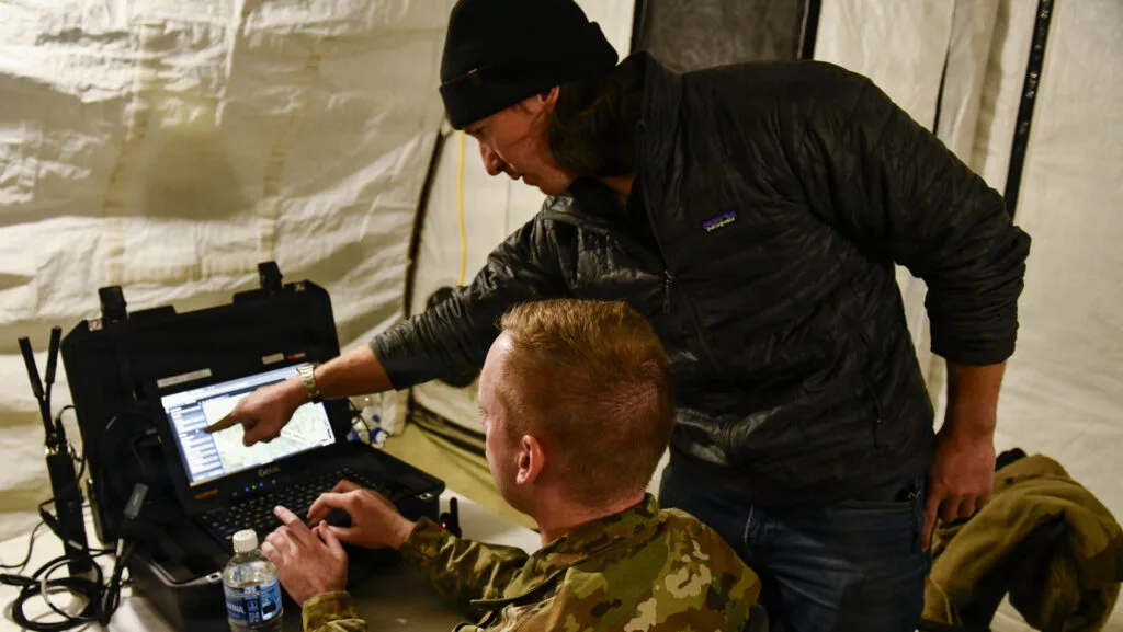 Dylan Driscoll, representante de Anduril, conversa con el aviador de primera clase Thomas Royce sobre la información que se introduce en Lattice. Este software forma parte de la función de mando y control del kit de despliegue rápido para UAVs del Comando Norte de los Estados Unidos. La imagen fue tomada durante un ejercicio en la Base de la Fuerza Aérea Minot, Dakota del Norte, el 23 de octubre de 2025.